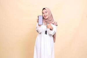 An Asian Muslim girl wearing a hijab smiles cheerfully, showing her cell phone gadget screen with a thumbs up gesture okay to create sales content. for advertising, technology, Eid and Ramadan photo