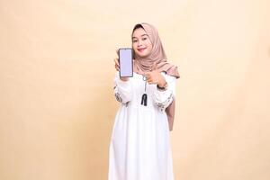 An adult Asian Muslim woman wearing a hijab smiles showing her cell phone gadget screen with a thumbs up gesture okay to create sales content. for advertising, technology, Eid and Ramadan photo