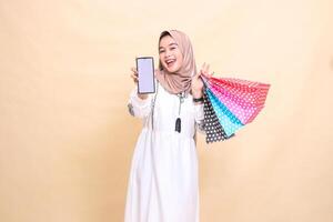 a young Asian Muslim woman wearing a hijab smiles happily, showing a cellphone screen and carrying a paper bag. for advertising, technology, Eid and Ramadan photo