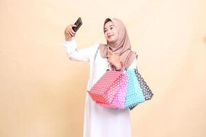 a young Asian Muslim woman wearing a hijab takes a selfie pointing at a cellphone camera and carrying a paperbag shopping bag. for advertising, culture, Eid and Ramadan photo
