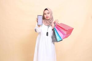 a young indonesia Muslim woman wearing a hijab is afraid to frown, showing a cellphone screen and carrying a paperbag shopping bag. for advertising, technology, Eid and Ramadan photo