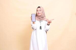 an adult indonesia Muslim woman wearing a hijab with a cheerful smile displays a cellphone screen gadget pointing left to create sales content. for advertising, technology, Eid and Ramadan photo