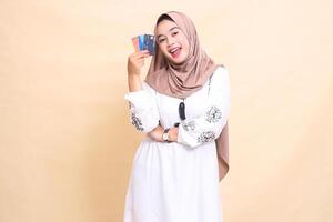 a beautiful indonesia Muslim woman wearing a hijab with a cheerful smile and her arms holding a debit credit card to her side for payment. used for advertising, technology, Eid and Ramadan photo