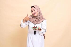 a beautiful Asian Muslim woman wearing a hijab smiles cheerfully with a thumbs up gesture okay good and brings some dates for the sahur menu and breaking the fast. used for health, Eid and Ramadan photo