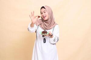 a beautiful Asian Muslim woman wearing a hijab with a cheerful smile and a wink took a date forward and brought some dates for the sahur menu and breaking the fast. used for health, Eid and Ramadan photo