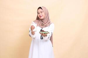 A beautiful Asian Muslim woman wearing a hijab smiles with a wink and brings a glass of milk and dates to the front while breaking the fast. used for advertising, health, Eid and Ramadan photo