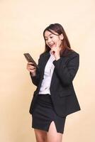 Beautiful formal Asian executive girl in suit standing cheerful expression getting ideas while operating online shopping gadget smartphone on yellow background for commercial, vacancies and business photo