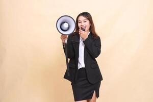 Beautiful formal Asian office woman in suit standing smiling shouting at center camera with mic using megaphone for promo broadcast announcement on yellow background for advertising and business photo