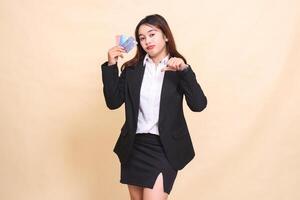 Beautiful formal indonesia office woman in suit standing sullenly holding debit credit card and thumbs up hand disappointed for transaction promotion on yellow background for commercial and business photo