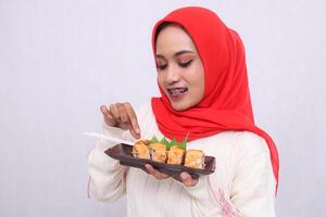 Asian woman in hijab cheerfully gestures to take a dish with her hand and both hands hold a plate containing sushi Japanese food. Beautiful Muslim women use culinary, food and lifestyle content photo