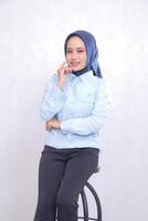 young Asian office woman wearing a hijab, blue shirt, sitting in a chair, smiling cheerfully, holding her cheeks with her arms crossed. Beautiful Muslim women use business, finance and fashion content photo