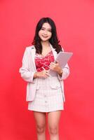 Portrait of a smiling young Asian Indonesian woman wearing a cream colored blazer carrying a tablet and pen. Isolated red background for content materials, banners, advertising. photo