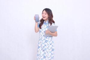 beautiful asian woman wearing blue chinese dress looking at cellphone happily holding tablet and looking at cellphone screen isolated white background photo