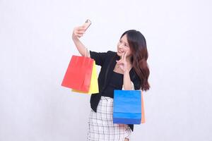 Beautiful young woman customer 20s formal office smiling happily while taking photo in front of cell phone with peace hands up while carrying shopping paper bag isolated white background