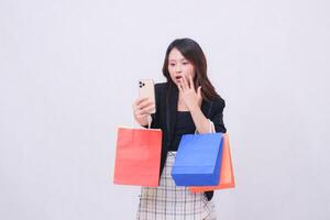 Beautiful Asian woman 20s formal office customer shocked looking at cellphone hand gesturing to shut up while carrying shopping paper bag isolated on white background photo