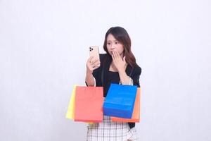 Beautiful Asian girl 20s formal office customer in shock looking at cellphone covering mouth while carrying shopping paper bag isolated on white background photo
