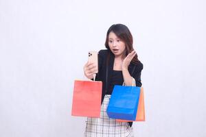 Beautiful Asian woman 20s formal office customer surprised looking at cellphone in left hand while carrying shopping paper bag isolated on white background photo