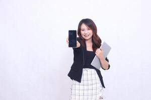 Beautiful Indonesian woman in her 20s, formal office, smiling, looking at a black cellphone gadget and carrying a tablet for promotion, isolated on a white background photo