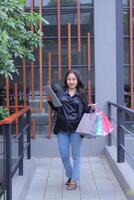 young Indonesian office girl is happy to get a discount price for an online order, looking at the camera, carrying a paper bag and laptop computer, to promote online content, lifestyle, catalogs photo