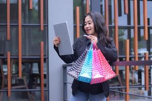 young Indonesian businesswoman happy to get discount on online order, standing against iron fence carrying paperbag and laptop computer, for promotion of online content, lifestyle, catalog photo