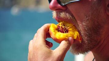 Man eating peach. Bearded man in sunglases eating peach on the beach. Happy man is smiling and resting at sea. Bearded guy enjoying fruits on summer vacation. Vegetarian and healthy lifestyle concept. video