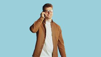 Young handsome caucasian man in glasses and casual clothes standing and starting to talk with a friend on a smartphone with a smiling face on a blue background in the studio video