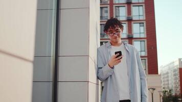Young asian curly man in casual clothes and glasses standing and looking at the smartphone with smiling face on a city street with multi-storey buildings as a backdrop video