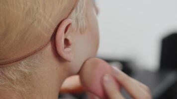 Young actor in a drag queen makeup applying the powder to his cheeks and forehead with a soft sponge in the dressing room during preparation for the performance close up, back view video