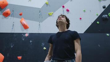 Confident young man with curly hair standing with arms crossed in front of an indoor climbing wall with colorful holds, showcasing strength and determination video