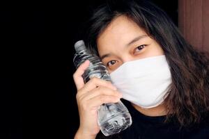 Asian Thai woman wearing a white cloth mask for prevent the Covid-19 or Corona virus and Air Pollution Value Pm 2.5 in Thailand and hand holding a bottled mineral water. Health and illnes Concept photo