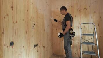 This shows a worker applying acrylic paint to a wooden wall in a new house. The worker is wearing work clothes and gloves. There is a ladder in the background. video