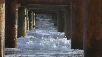 Waves with white foam break against large iron and rusty columns. CREATIVE. Sea waves are raging under the pier near the shore video