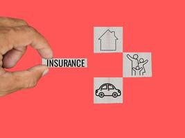 The hand is gripping a wooden block with the word insurance written on it, along with cubes representing the concepts of home, family, and car. featuring a pale red backdrop photo