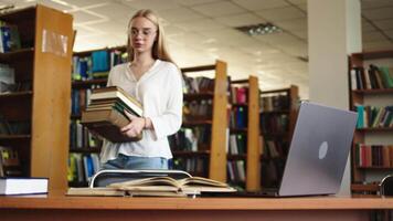 atraente multiétnico aluna fazer uma haste classe dever de casa dentro uma livro didático, enquanto pesquisando questionário respostas em uma computador portátil computador. jovem Loiras fêmea estudando dentro uma público biblioteca com clássico interior video