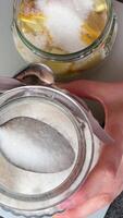 top view of hands pouring granulated sugar on lemon slices in glass jar. Unrecognisable person, slow-mo zoom out. Close-up video