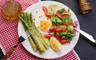 Round plate with cooked asparagus, fried egg, avocado and fresh vegetable salad on the table, top view photo
