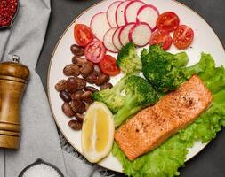 Healthy lunch with grilled salmon on green lettuce, next to vegetables, tomatoes, radishes, broccoli and a portion of beans. photo
