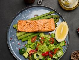 Healthy lunch with grilled salmon with asparagus and fresh tomato and cucumber salad, a row of lemon slice. photo