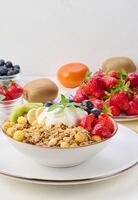 Granola with strawberries, kiwi, banana and blueberries in a round plate topped with yogurt on the table. photo