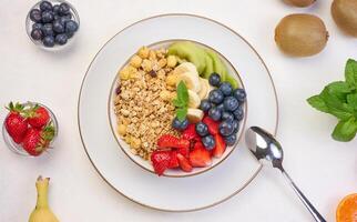 Granola with strawberries, kiwi, banana and blueberries in a round plate on a white table. photo