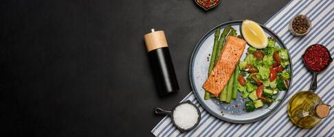 Healthy lunch with grilled trout with asparagus and fresh tomato and cucumber salad, a lemon slice. Food on a blue round plate, top view photo