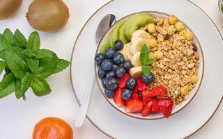 Granola with strawberries, kiwi, banana and blueberries in a round plate on a white table. Healthy and tasty food photo