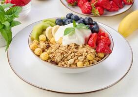 Granola with strawberries, kiwi, banana and blueberries in a round plate topped with yogurt on the table. photo