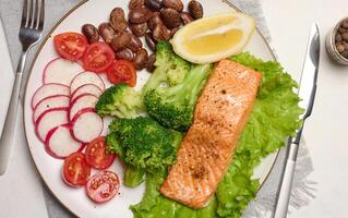 Healthy lunch with grilled salmon on green lettuce, next to vegetables, tomatoes, radishes, broccoli and a portion of beans. photo