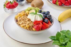 Granola with strawberries, kiwi, banana and blueberries in a round plate on a white table. photo