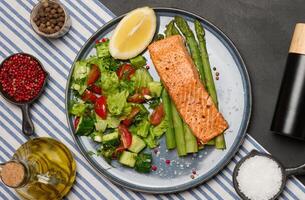 Healthy lunch with grilled salmon with asparagus and fresh tomato and cucumber salad, a row of lemon slice. photo