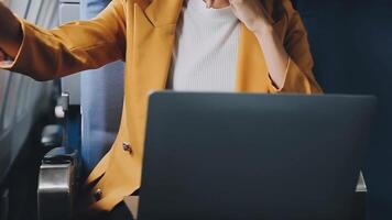 Attractive asian female passenger of airplane sitting in comfortable seat listening music in earphones while working at modern laptop computer with mock up area using wireless connection on board. video