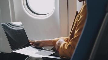 Attractive asian female passenger of airplane sitting in comfortable seat listening music in earphones while working at modern laptop computer with mock up area using wireless connection on board. video
