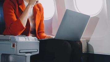 Attractive asian female passenger of airplane sitting in comfortable seat listening music in earphones while working at modern laptop computer with mock up area using wireless connection on board. video