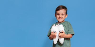A smiling boy with healthy teeth holds a plush tooth in his hands on a blue isolated background. Oral hygiene. Pediatric dentistry. Prevention of caries. A place for your text. photo